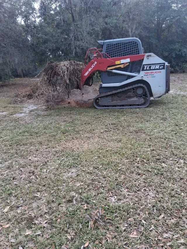A red and white Takeuchi track loader pushing a large pile of brush across a grassy, wooded lot.