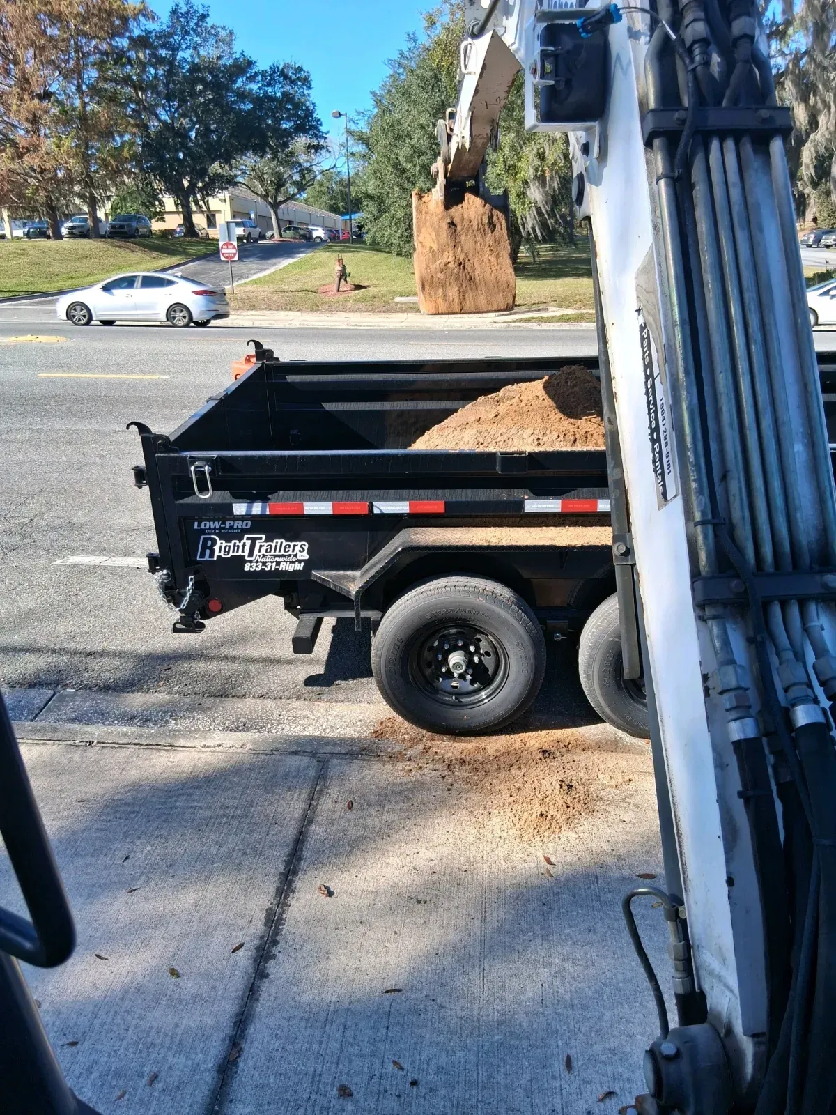 An excavator bucket dumping dirt into a black dump trailer parked on a road with a white car passing by.