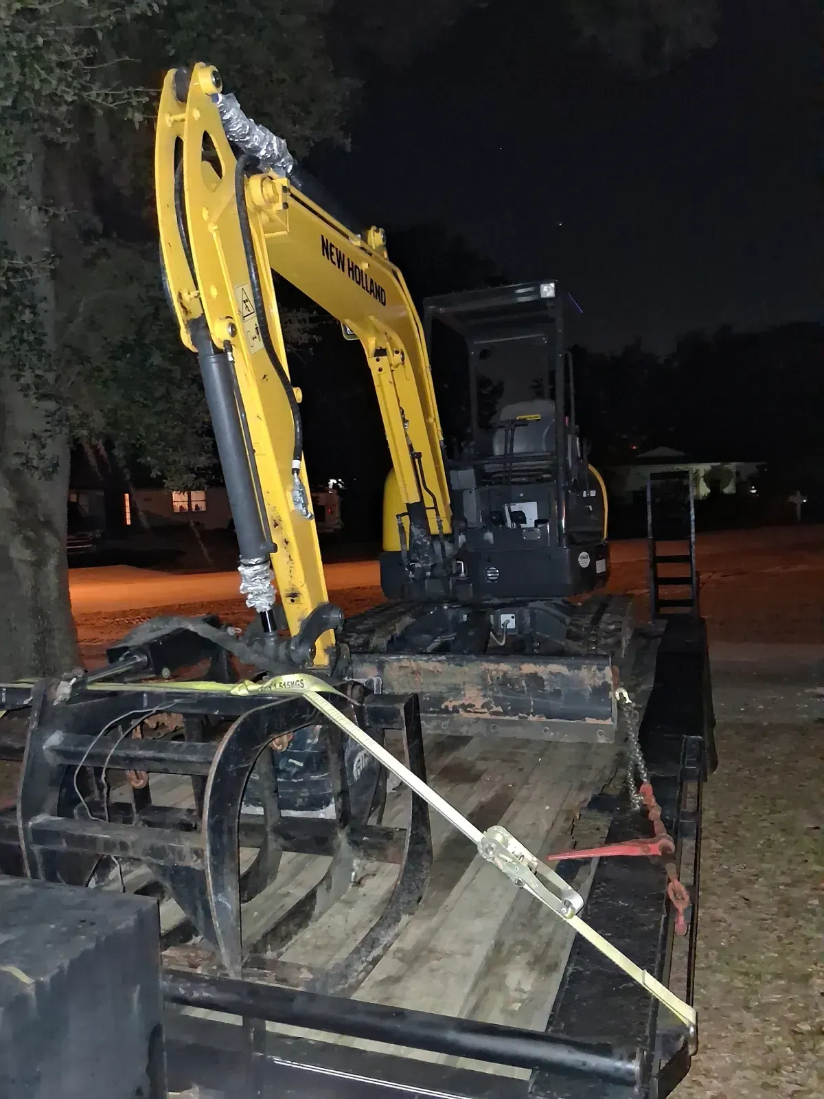 A yellow excavator with a grapple attachment sits secured on a flatbed trailer at night.