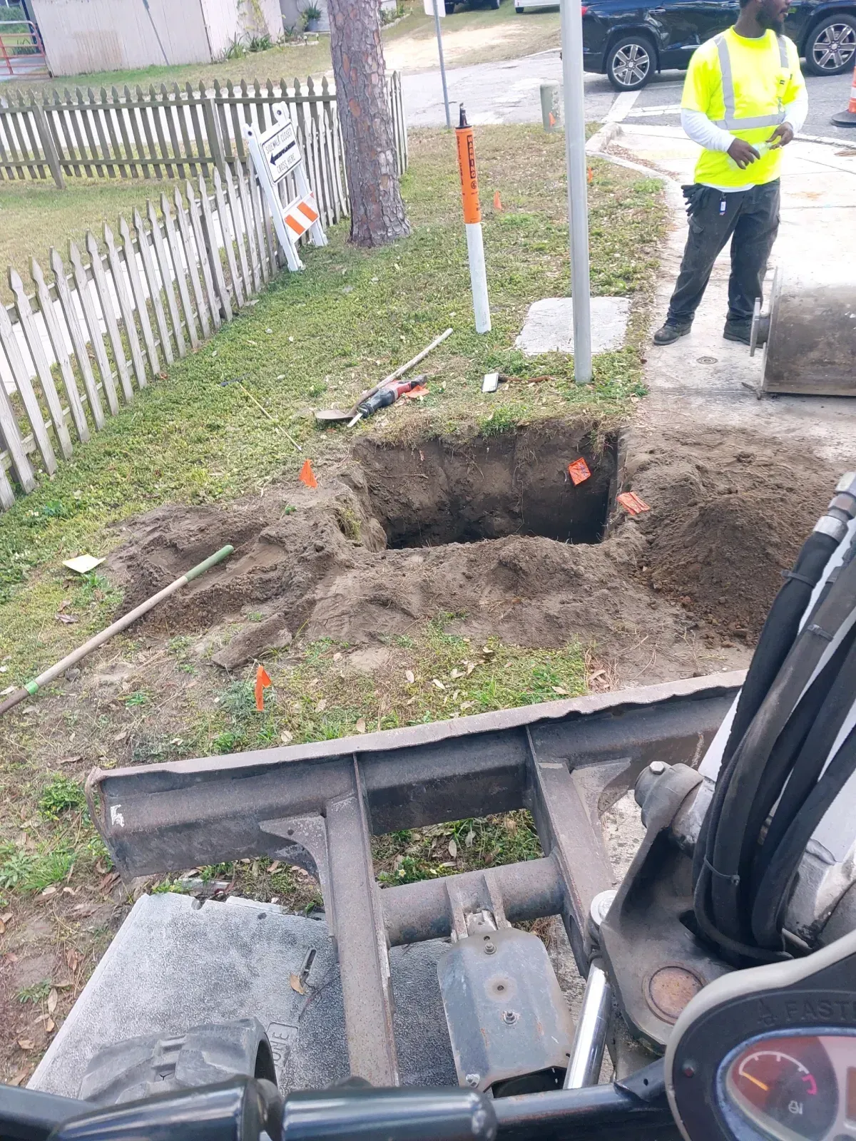 A construction site with a worker in high-visibility gear standing near a dug trench by a residential fence.