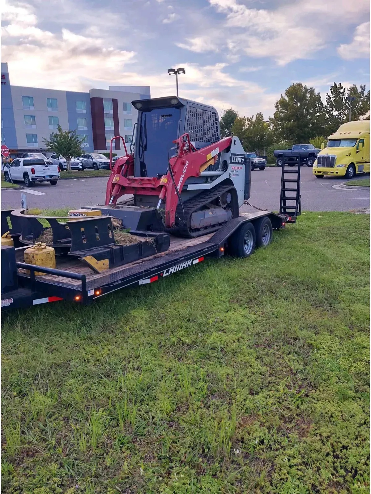 A red and white skid-steer loader sits secured on a flatbed trailer parked on grass near a parking lot.