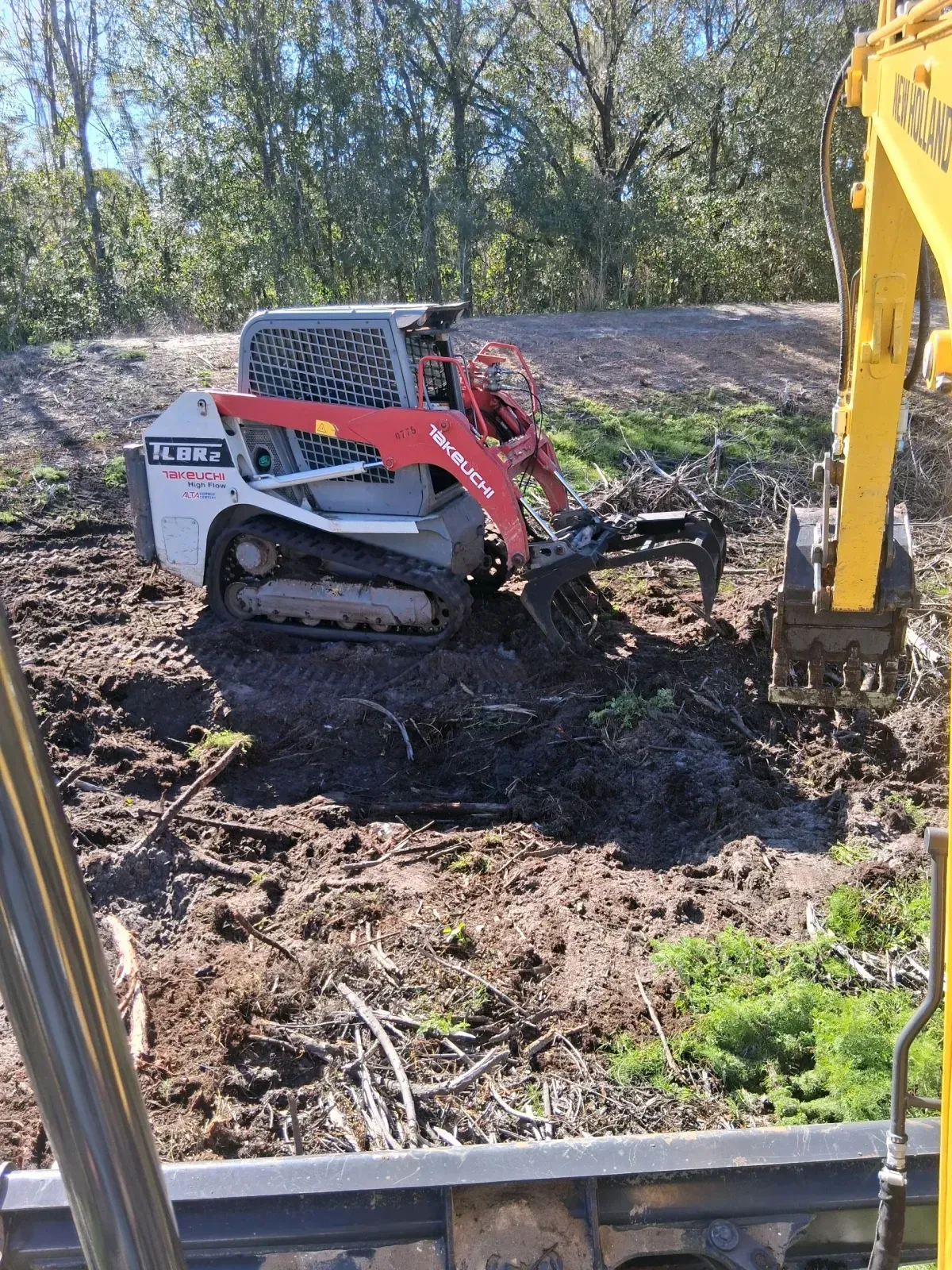 A white and red Takeuchi track loader clearing debris on a muddy construction site, viewed from another machine.