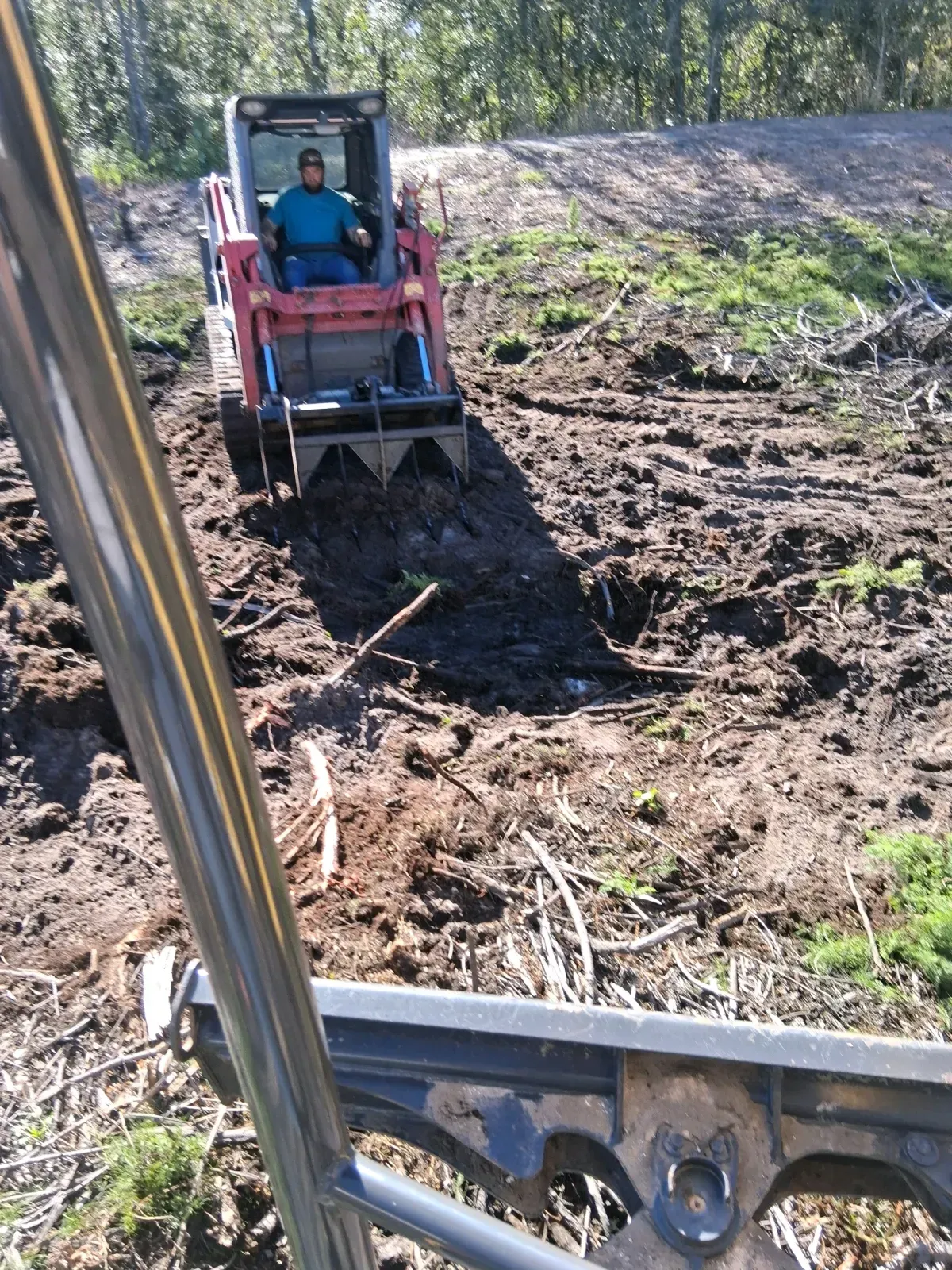 An operator drives a red skid steer with a forestry grapple attachment across a muddy, cleared landscape.