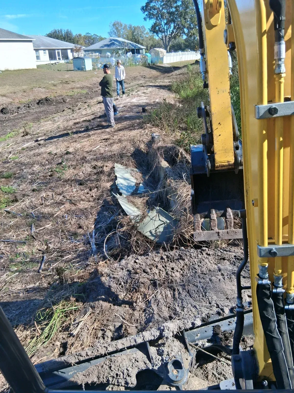 An excavator sits on a dirt path with two people standing nearby in a suburban area with homes in the background.