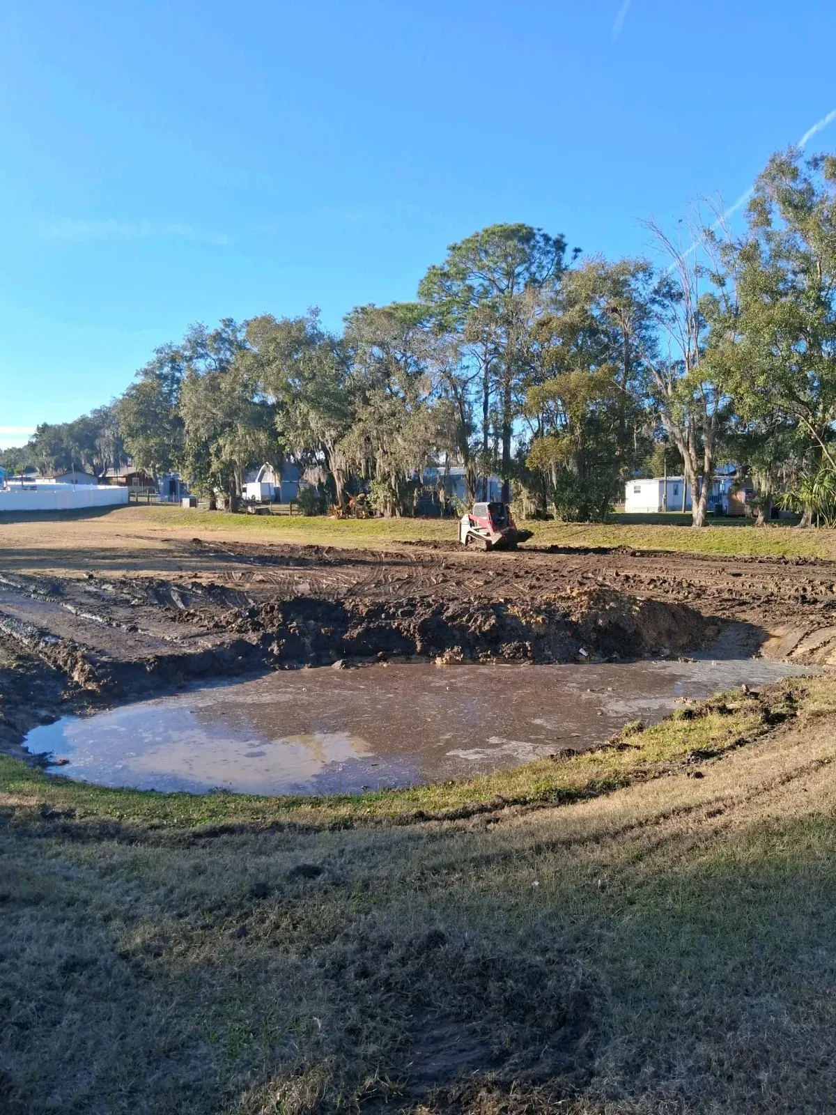 A skid steer loader works on a muddy, excavated lot with a small pool of water in the foreground under a clear blue sky.