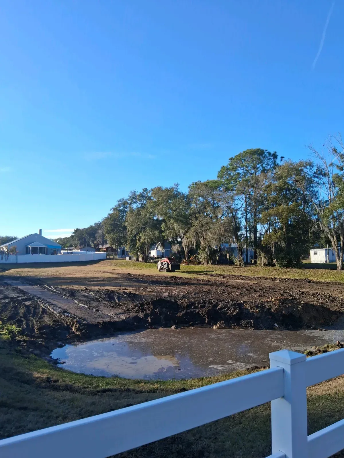 A muddy construction lot with a small puddle in the foreground, framed by a white fence and distant trees under blue sky.