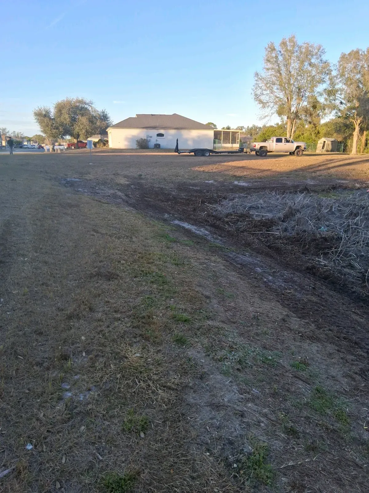 A flat, grassy field with a white house in the distance, parked trucks, and a patch of dark, tilled soil in the foreground.