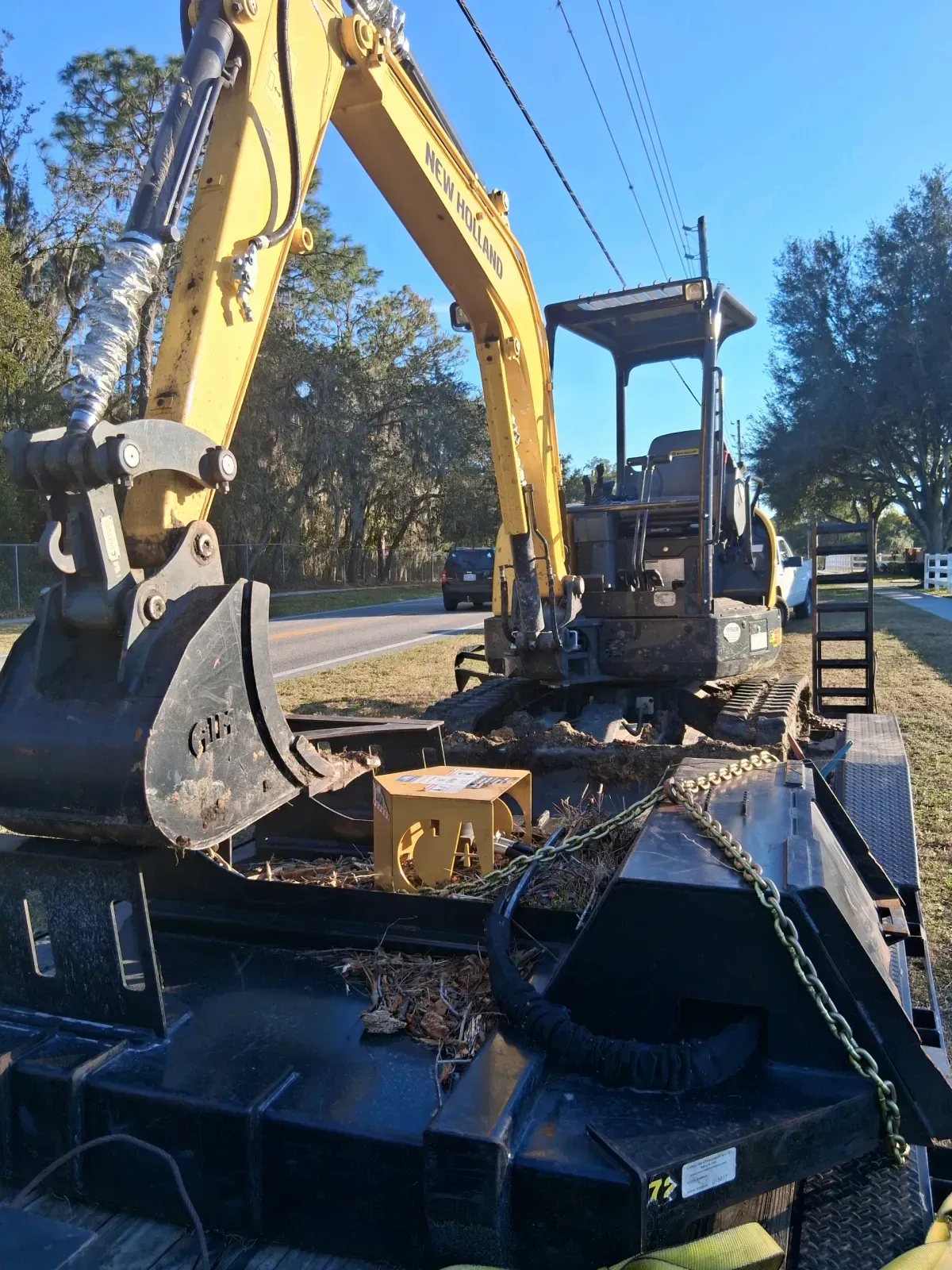 A yellow excavator sits on a flatbed trailer on a roadside, with a clear blue sky in the background.