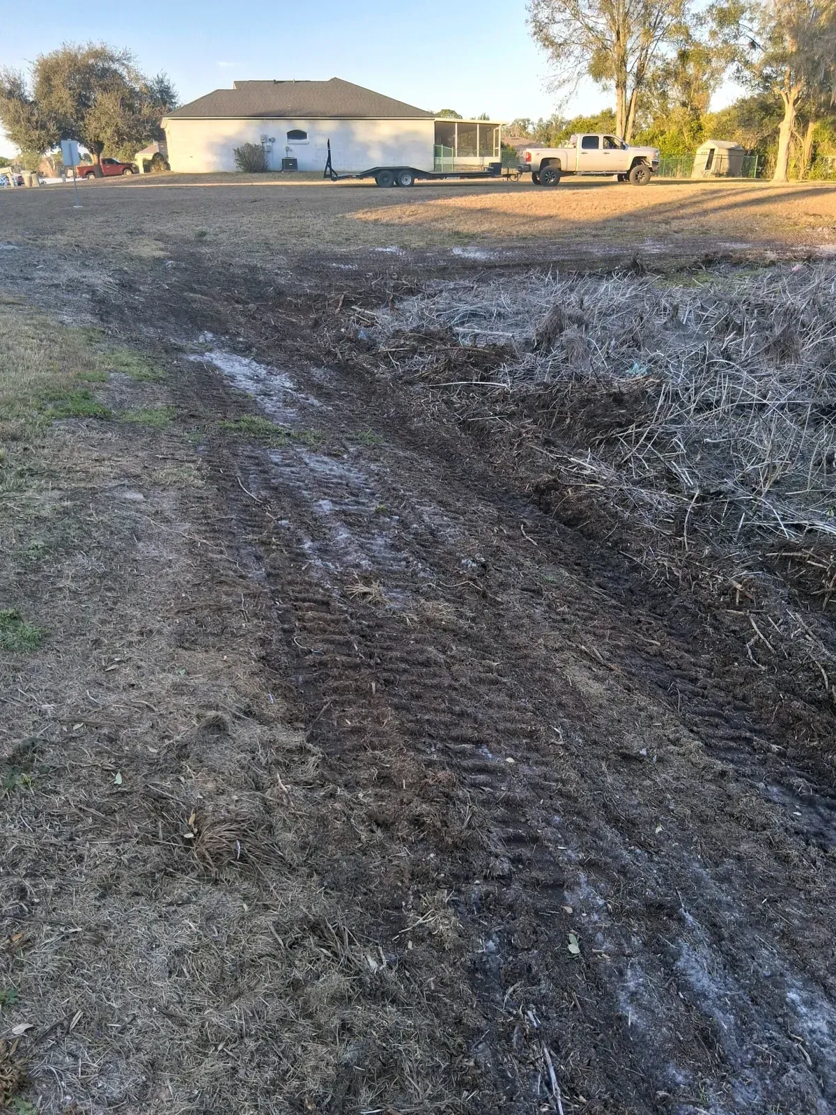 Deep tire tracks cut through mud in a grassy field in front of a house, trailer, and truck.