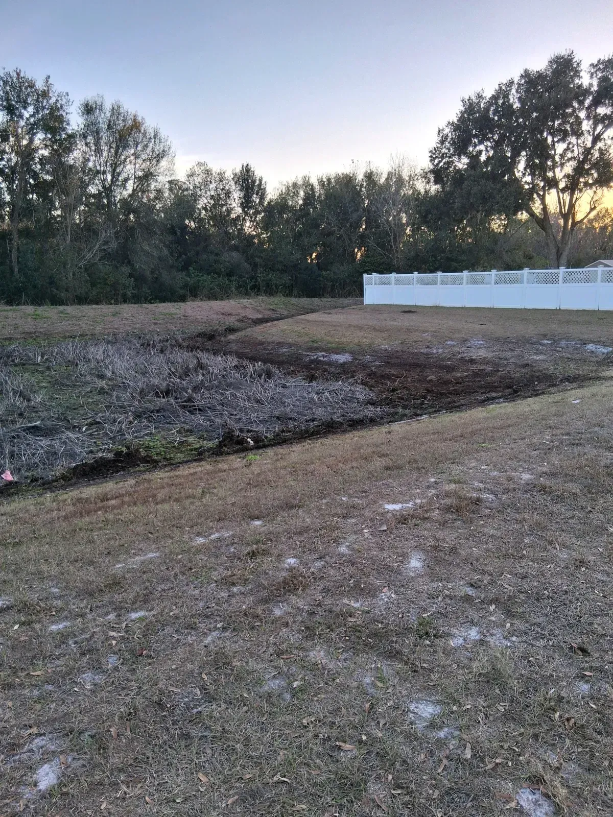 A view of a grassy backyard showing a cleared, dark patch of earth leading toward a white fence and wooded area.