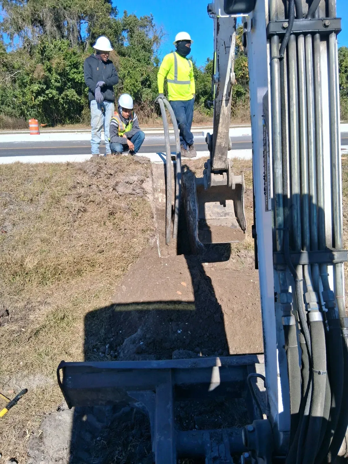 Three workers in high-visibility gear stand near a construction excavator at an outdoor worksite.