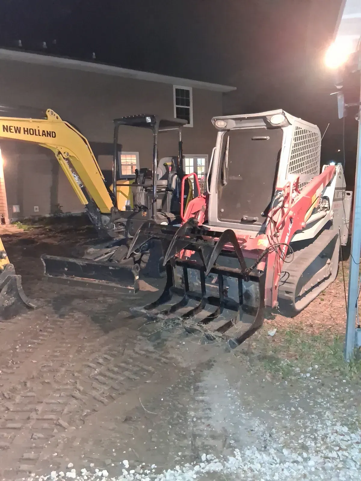 A yellow New Holland excavator and a white track loader with a metal rake attachment parked on a dirt lot at night.