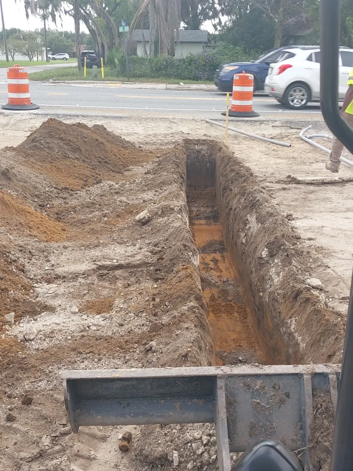 A narrow trench dug in the ground at a construction site, with orange traffic barrels and vehicles on a nearby road.