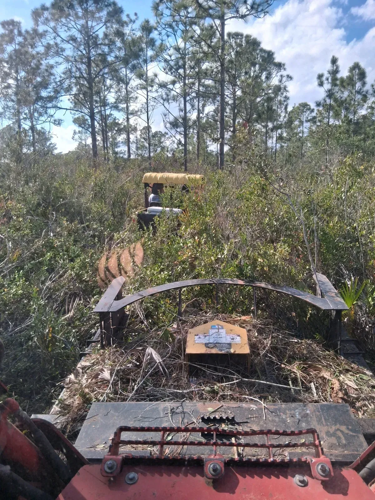 A view from the cockpit of a brush mower driving through thick woods toward a parked UTV under tall pine trees.