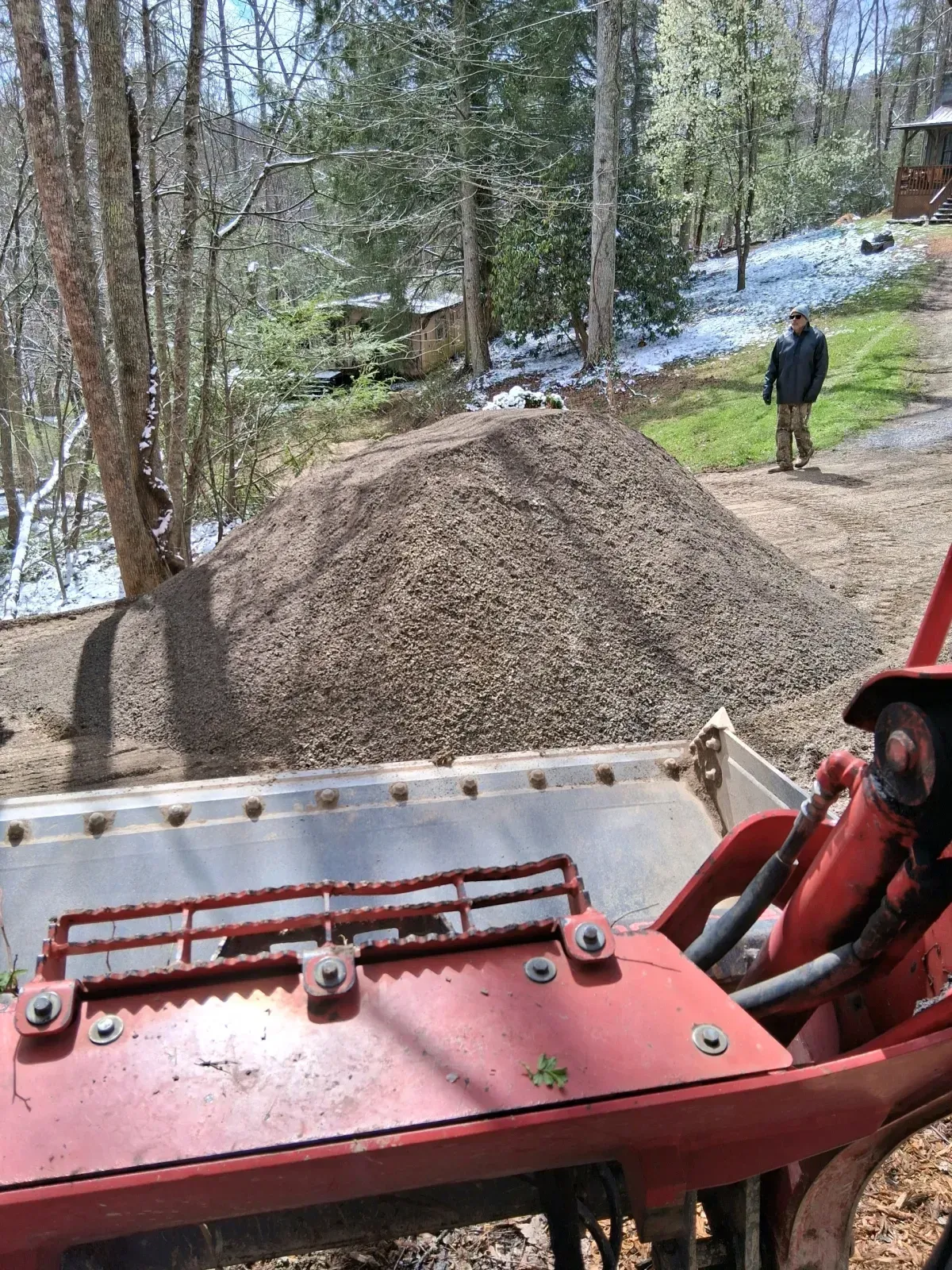 A view from a red tractor's cabin looking toward a large pile of gravel in a wooded area with a person standing nearby.