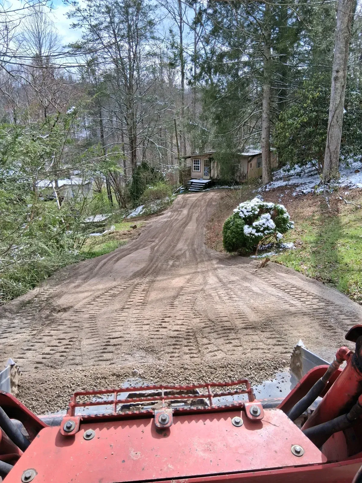 View from inside a machine looking down a steep, freshly graded dirt driveway toward a house nestled in a snowy forest.