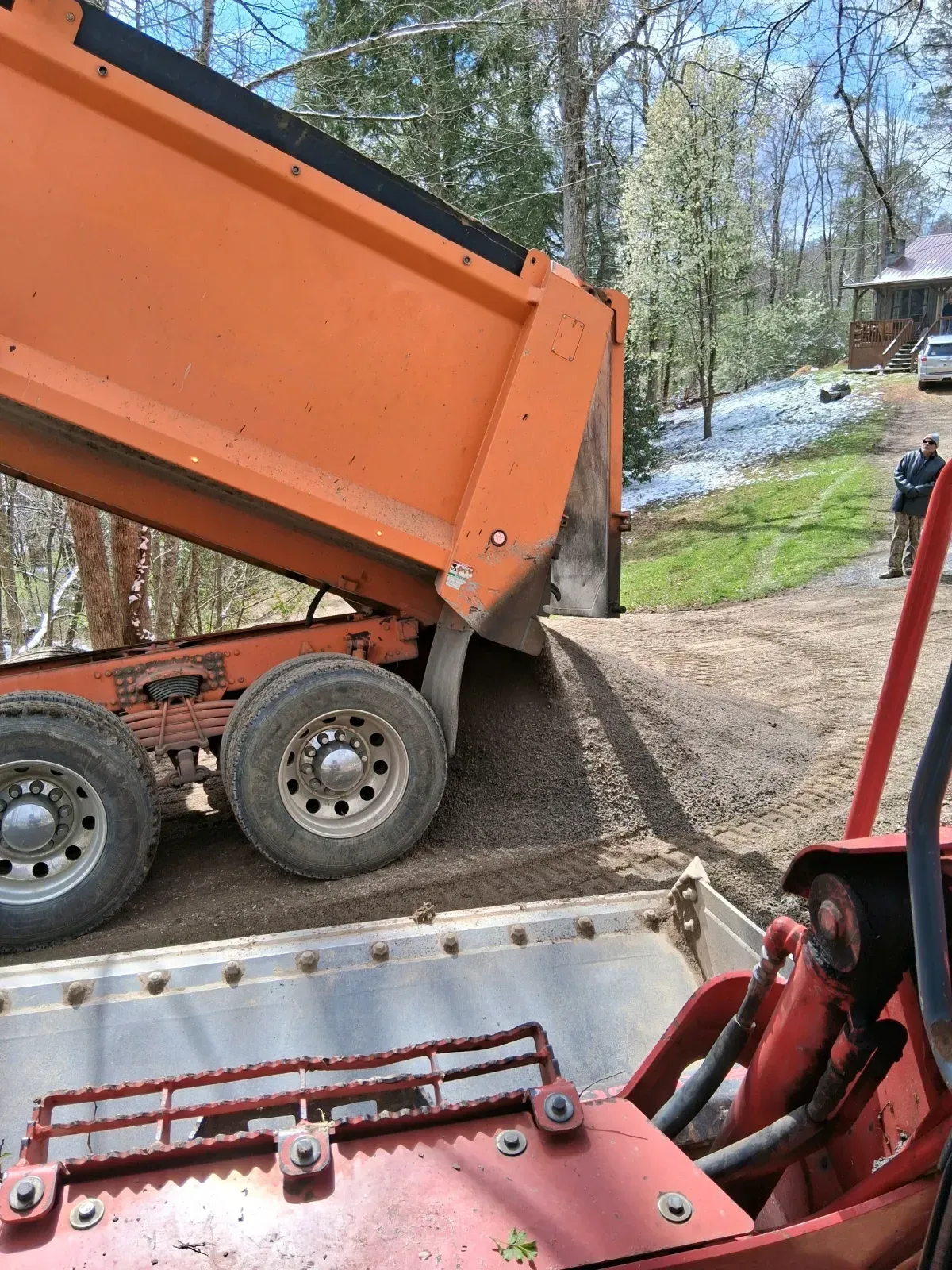 An orange dump truck deposits gravel onto a dirt driveway, viewed from the cab of a red tractor.