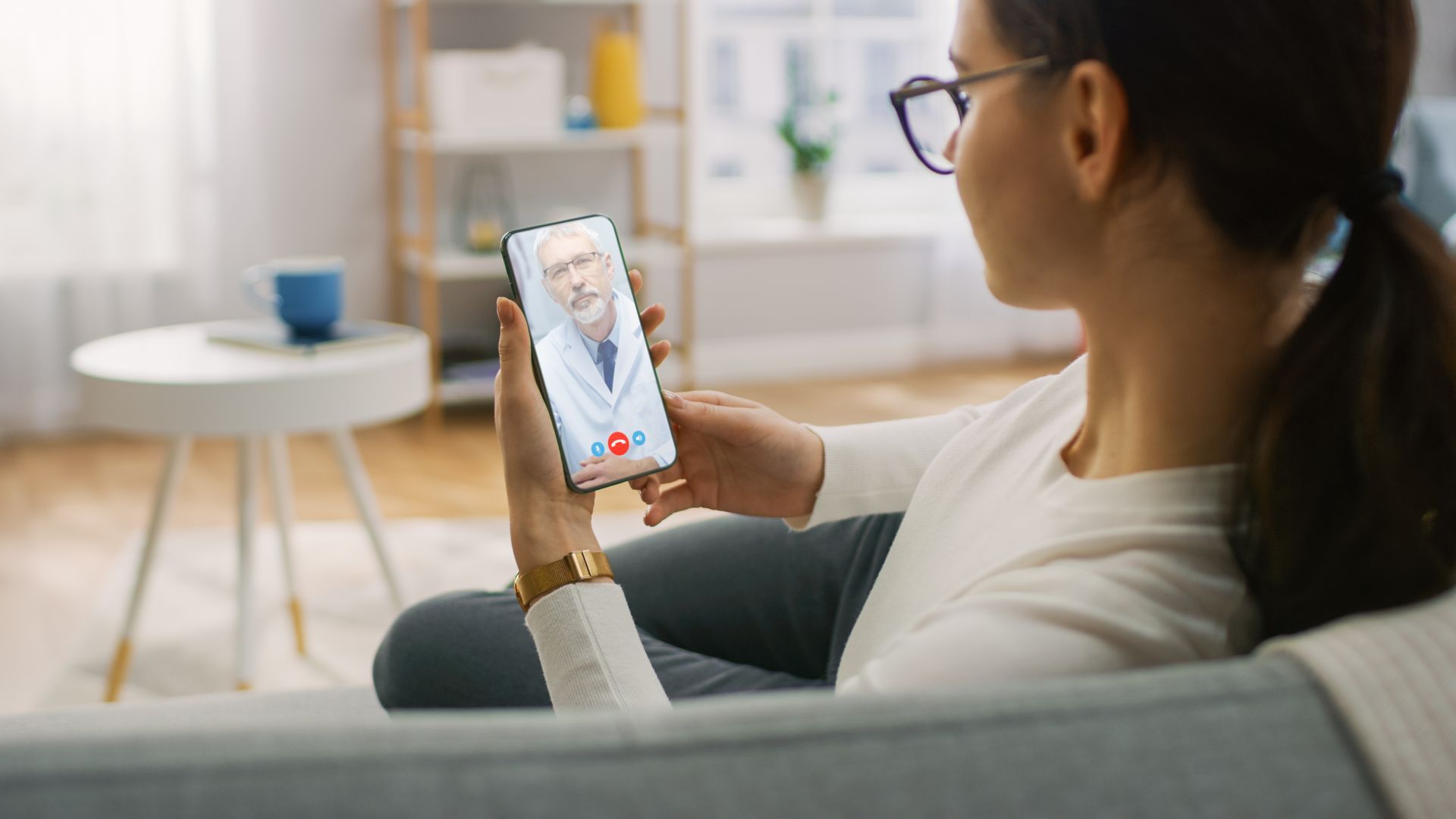 A woman is sitting on a couch holding a cell phone with a doctor on it.