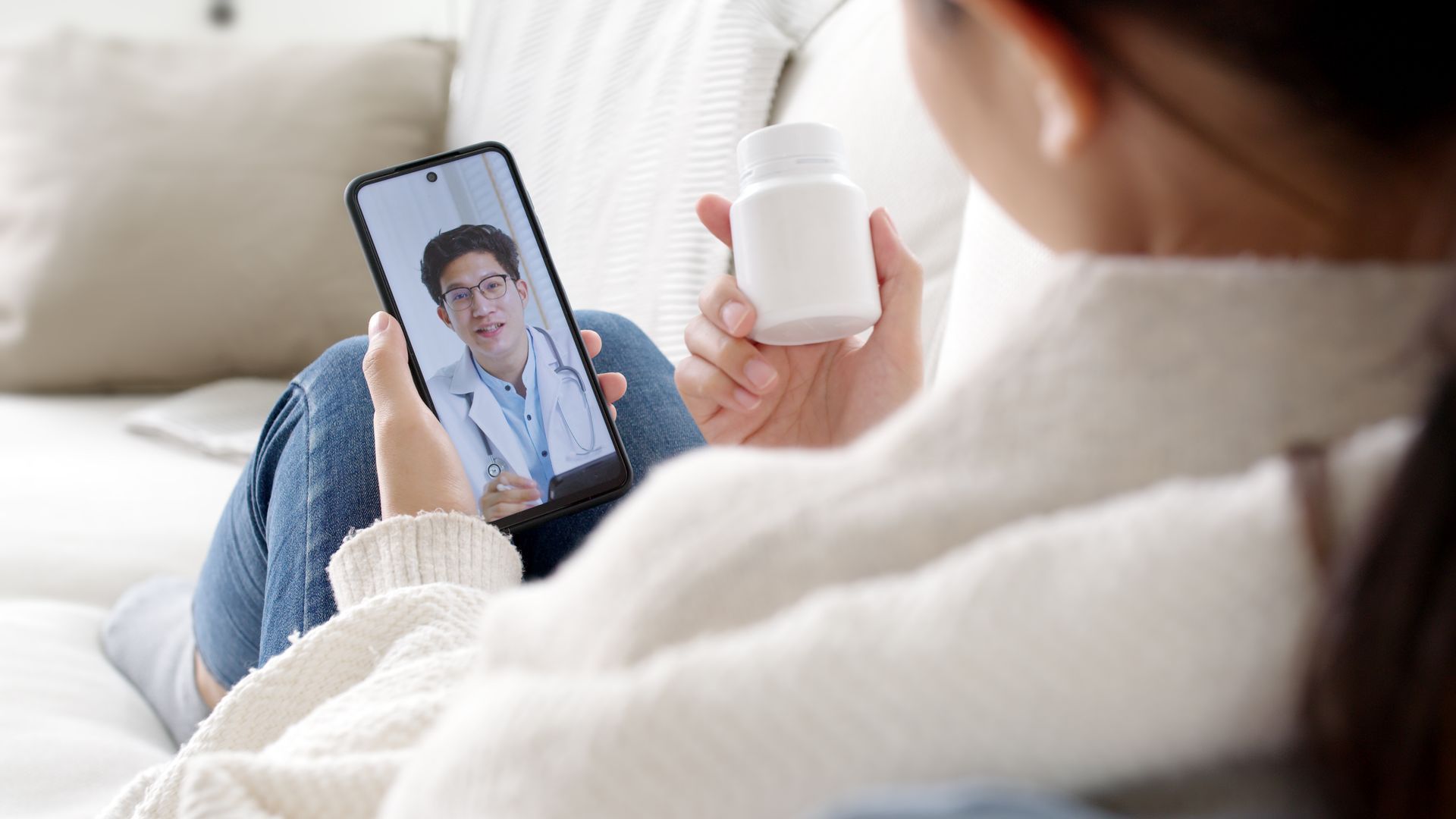 A woman is sitting on a couch holding a bottle of pills and talking to a doctor on her phone.