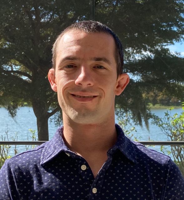 A man in a blue polka dot shirt is smiling in front of a lake.