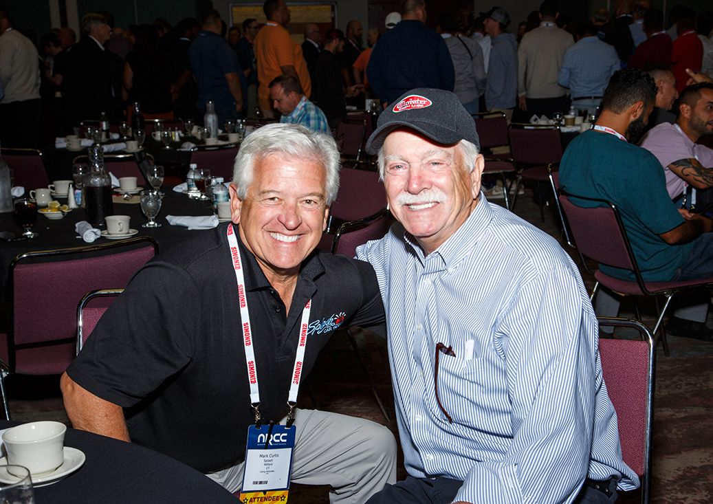 Two smiling men sitting together at a banquet table in a crowded event hall