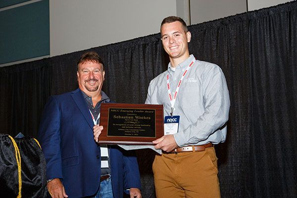 Two men holding a plaque at a conference, standing in front of a black curtain.