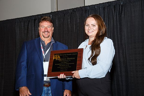 Two people holding a plaque in front of black curtains, smiling at an awards ceremony.