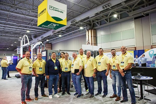 Group of exhibitors in yellow shirts posing at a trade show booth under bright convention hall lights
