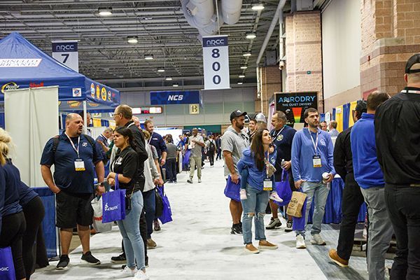 People lining an indoor convention hall aisle, many in blue shirts and carrying bags