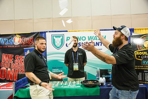 Three men at a trade show booth with banners, one speaking and gesturing beside display tables