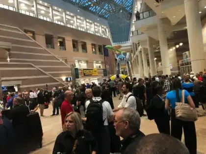 Crowded indoor atrium with people walking, escalators, and a large glass ceiling overhead