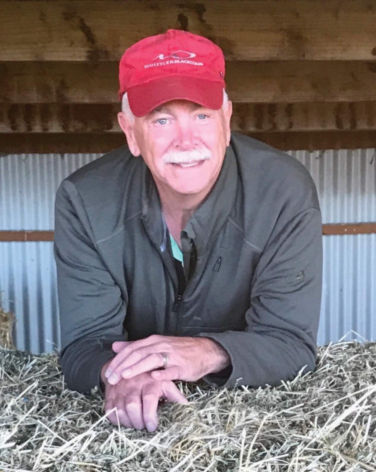 Smiling man in a red cap and dark jacket leaning on hay inside a barn