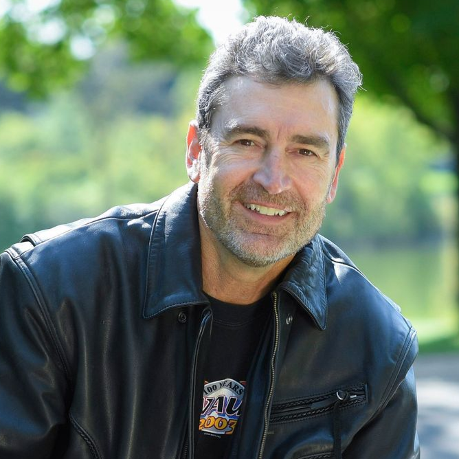 Man smiling on a motorcycle in a sunny park, wearing a black jacket and jeans.