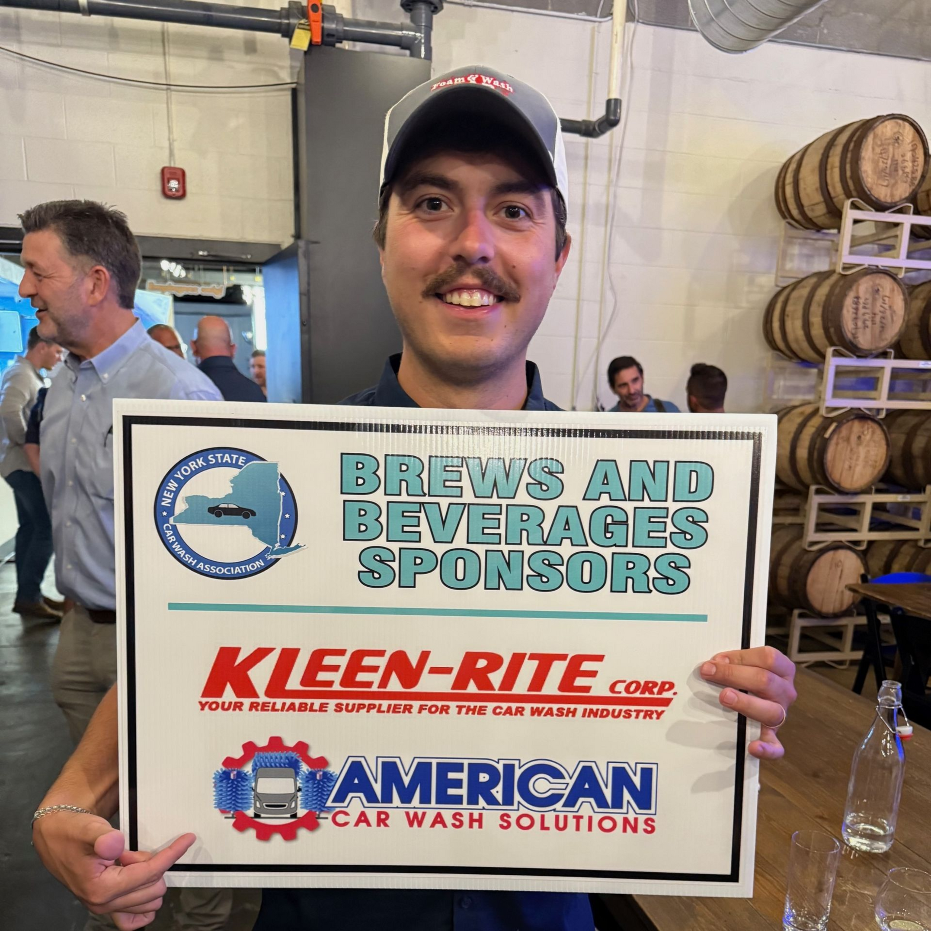 Man holding a sponsor sign with brewery and car wash logos in an indoor event space