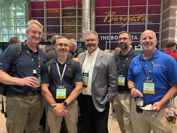 Five men posing together at a conference booth with drinks and lanyards, smiling in front of a Bordeaux sign