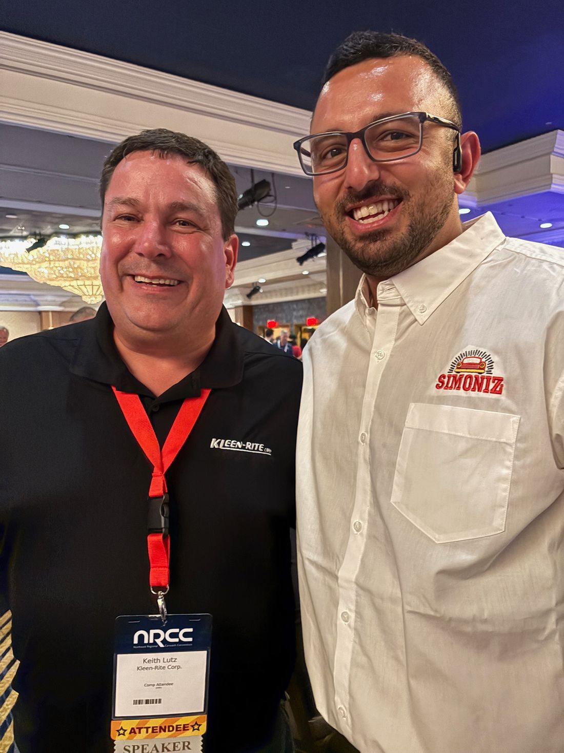 Two men smiling at an indoor event, one in a black polo and one in a white shirt with a red logo badge