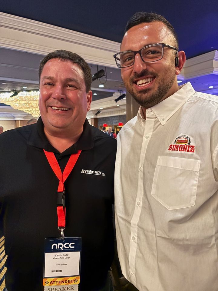Two men smiling at an indoor event, one in a black polo and one in a white shirt with a red logo badge