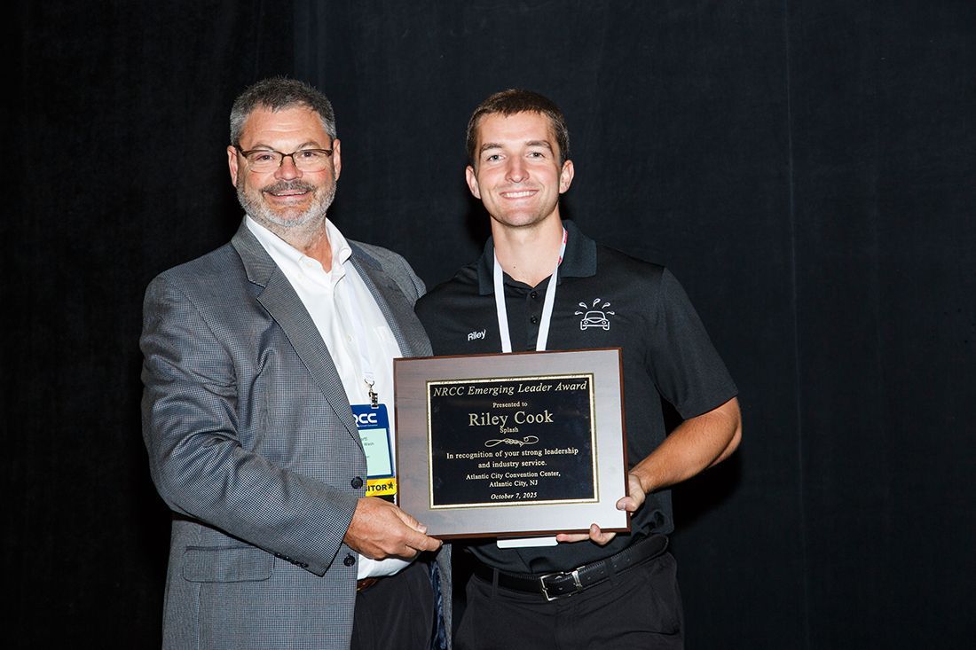 Two men in suits hold an award plaque on a dark stage.