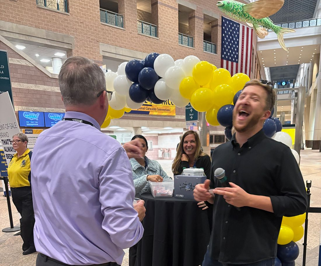 Man speaking into a microphone at a lively indoor event with blue, white, and yellow balloon decorations and onlookers
