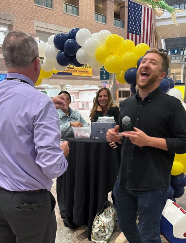 SDtuart and Chris Man speaking into a microphone at an indoor event with black, white, and yellow balloons behind him.