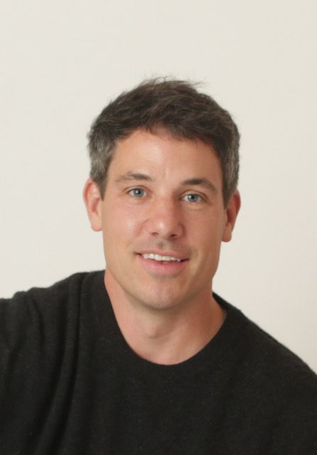Man smiling on a motorcycle in a sunny park, wearing a black jacket and jeans.
