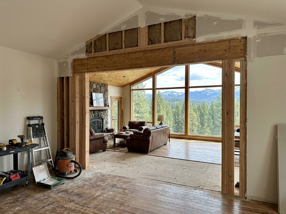 Open-plan cabin interior with exposed wood beams and large windows overlooking forest and mountains.