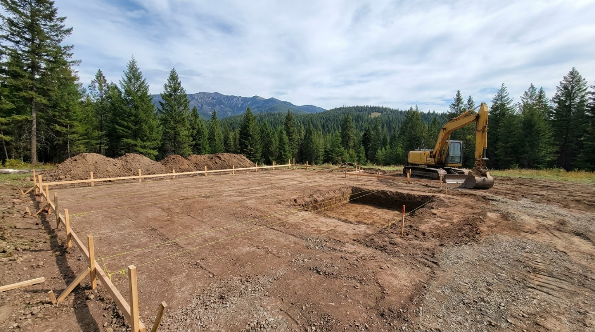 Cleared dirt lot with stakes, a yellow excavator, and evergreen trees under a cloudy sky