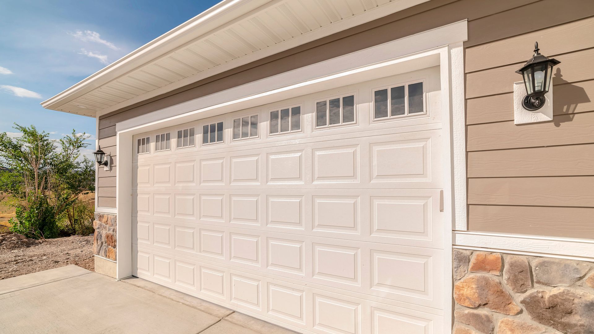 A white garage door with rectangular windows, attached to a house with beige siding and stone accents under a blue sky.