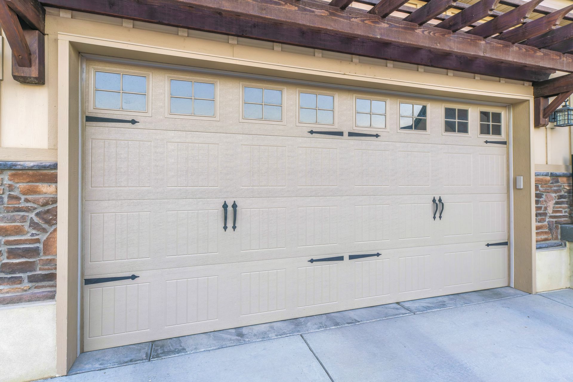 A tan garage door with decorative black hinges, handles, and small window panes in a stone-accented home exterior. A tan garage door with decorative black hinges, handles, and small window panes in a stone-accented home exterior.