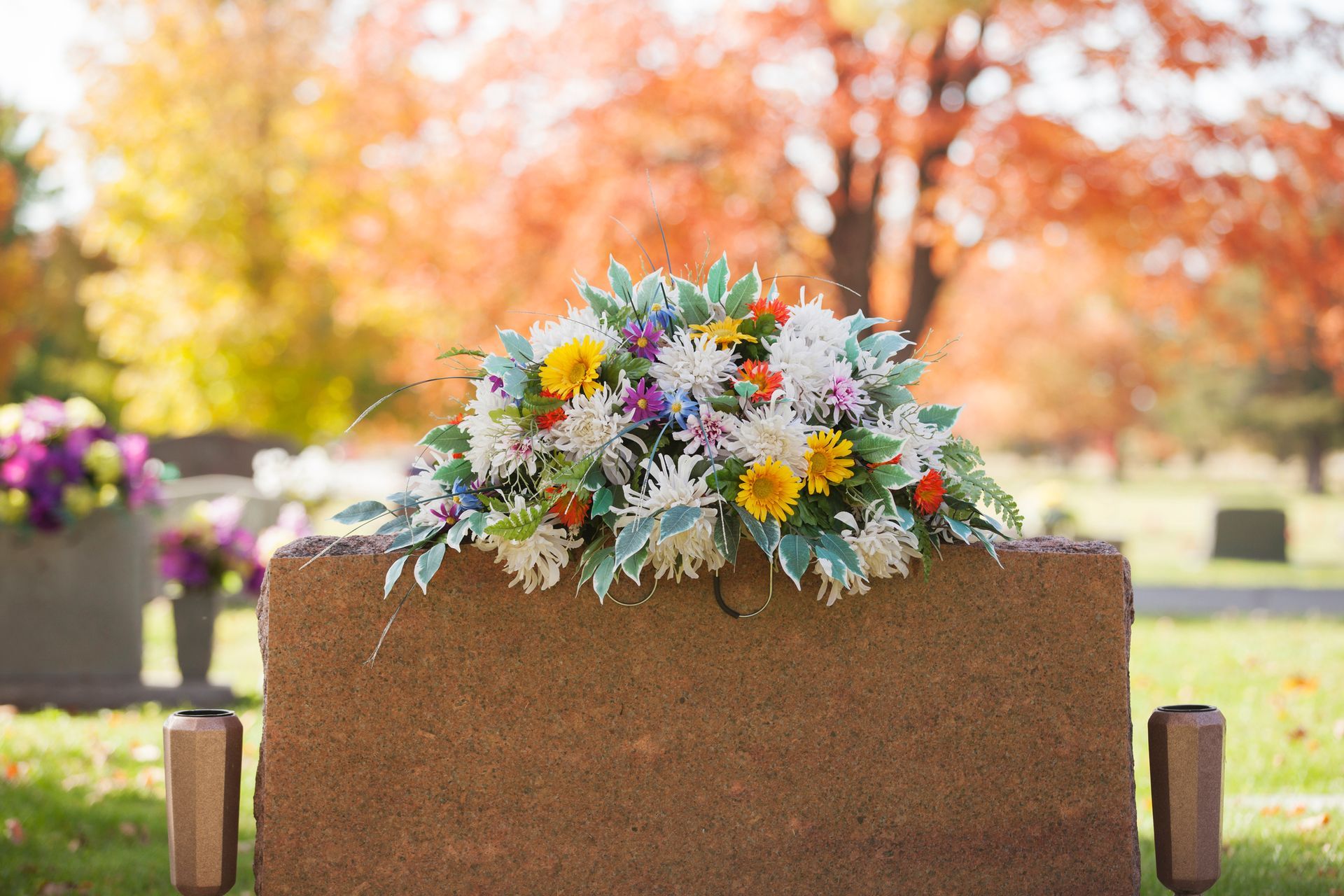 Grave with colorful flower arrangement in a cemetery with fall foliage in the background.