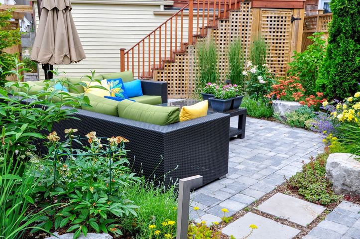 Backyard patio with wicker sofa, colorful cushions, potted flowers, and stone walkway beside stairs
