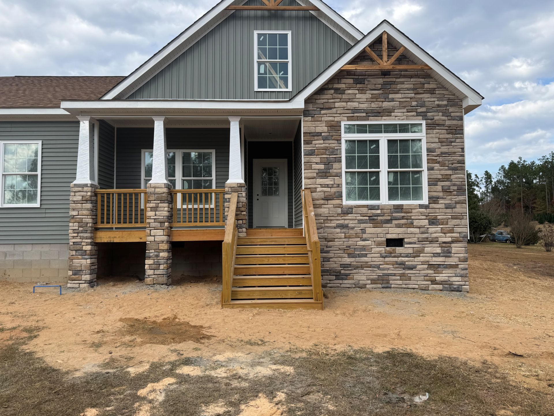 Front view of a gray house with a stone facade, white porch columns, and wooden steps over bare dirt.