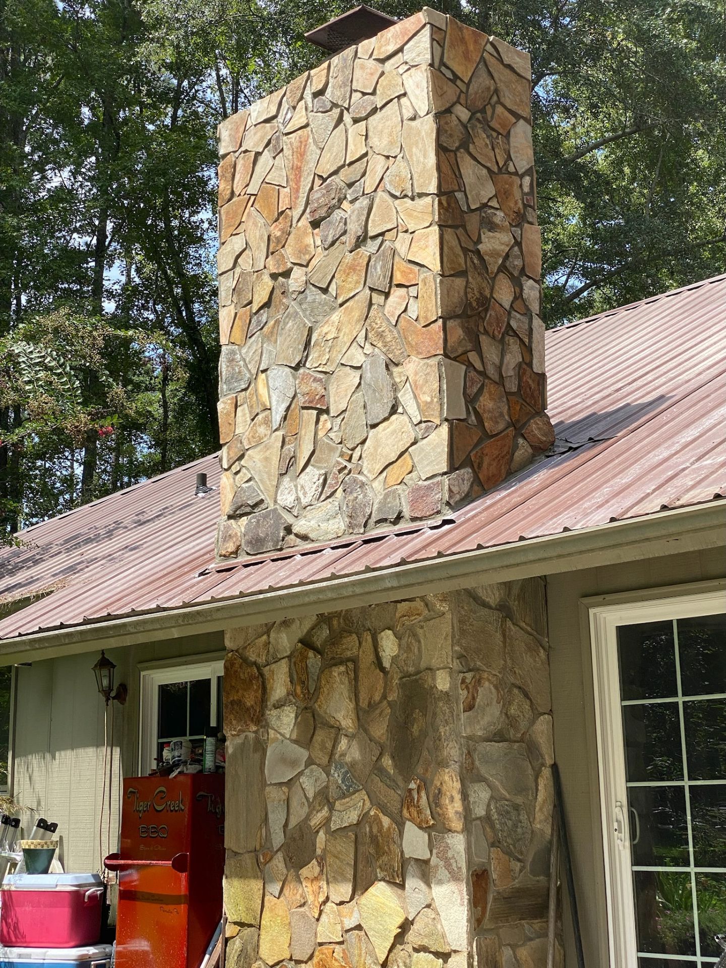 Stone chimney on a house with a red metal roof and trees in the background