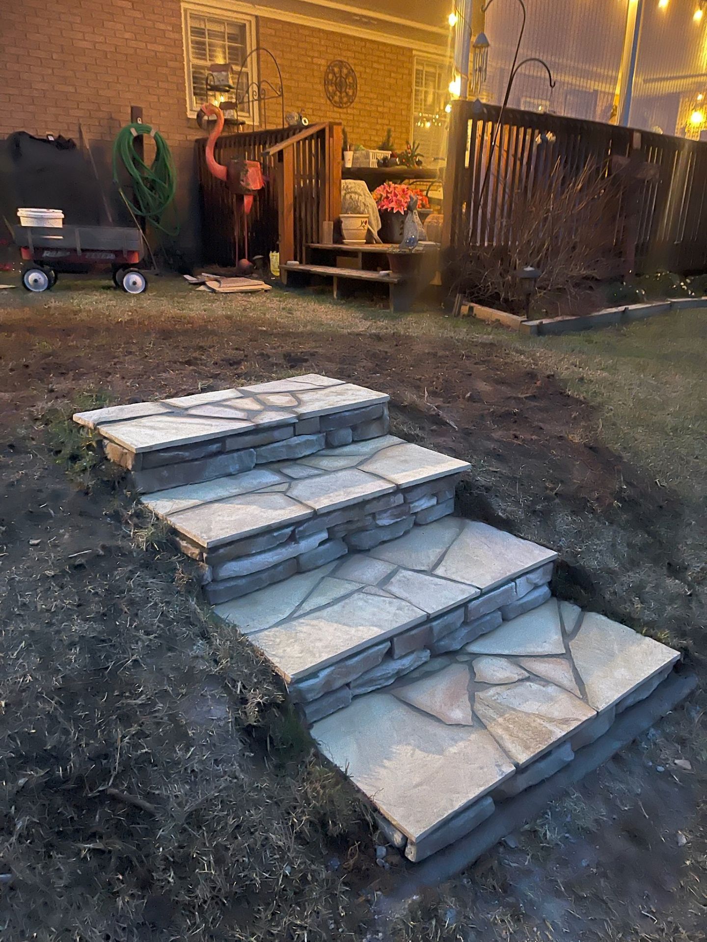 Stone garden steps leading to a backyard patio at dusk with a fence and warm porch lights